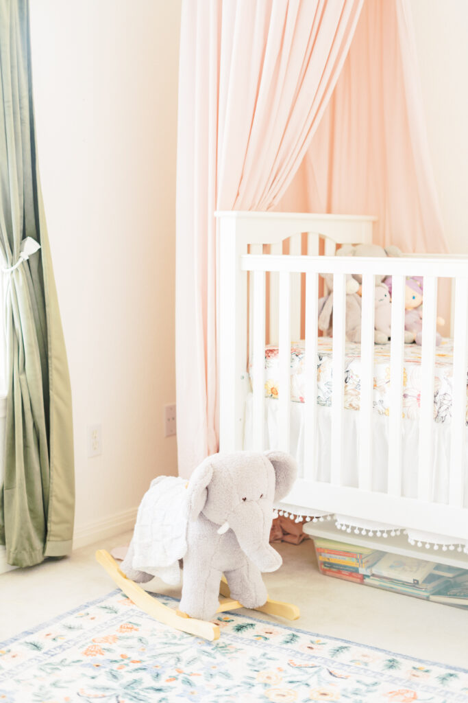 A nursery fit for a girl showcases a white crib with a pink canopy, a soft pink blanket, and stuffed animals inside. Nearby, a gray elephant rocker rests on a colorful floral rug. Light streams through green curtains, creating an airy and serene atmosphere perfect for lifestyle newborn sessions.