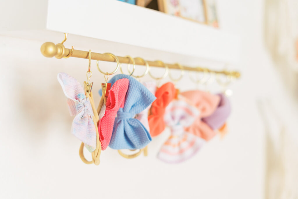 A row of colorful hair bows, including blue, pink, and orange, hangs from golden rings on a wall-mounted rod in a light and airy nursery setting.