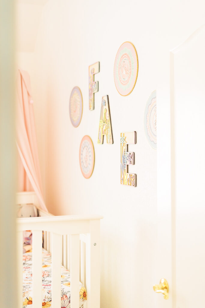 A nursery room with white walls showcases vibrant circular and letter decorations spelling the name "FAE" above a white crib. The light and airy space, adorned with nursery decor including a pink canopy over the patterned mattress, is worthy of an Austin newborn photographer. The room feels bright and welcoming.