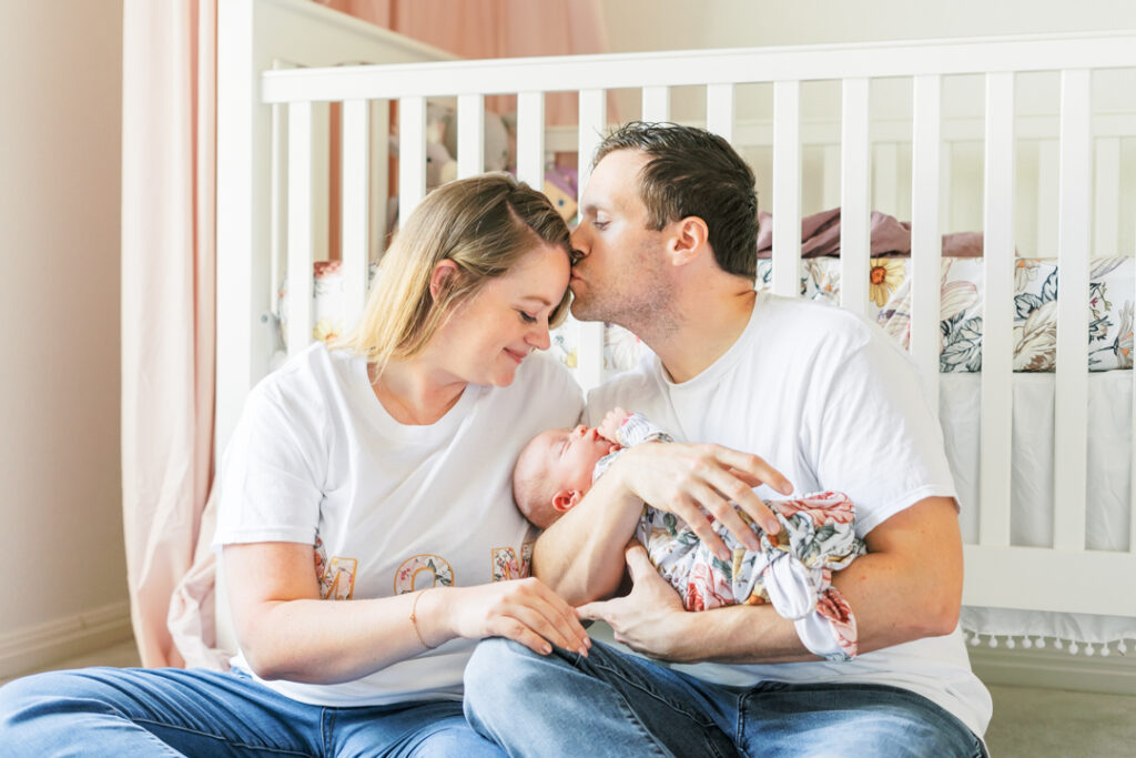 A mother and father lean against the front side of a white crib in a nursery filled with natural light. The father holds his newborn baby girl while kissing the mother on the forehead. The mother is peacefully looking down at her baby while smiling. The tender and timeless moment is beautifully captured by an Austin newborn photographer.