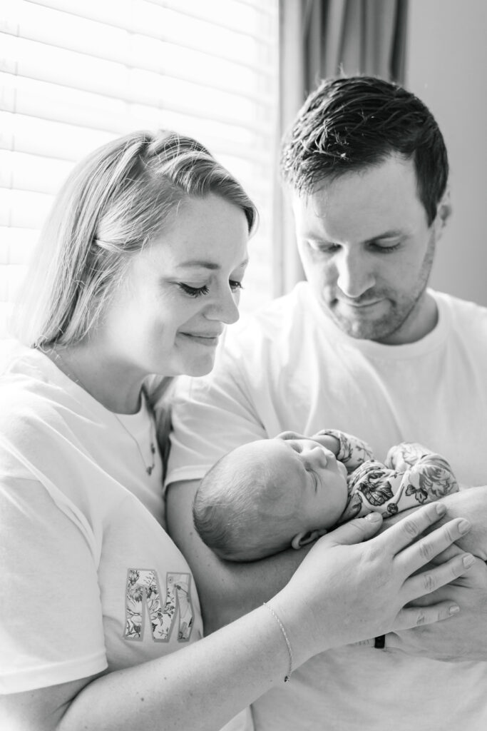 A mother and father look down at their newborn baby while standing with their back against the nursery's window. The father holds the newborn while the mother places her hand on the fathers arm and smiles. The black and white photo is beautifully captured by an Austin newborn photographer.