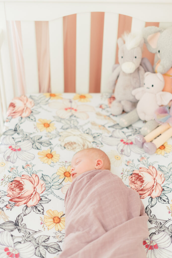 A newborn baby girl is swaddled in a mauve muslin blanket and peacefully sleeping on a floral fitted shit in a white crib. Above her head, to the right, are stuffed animals. There is a pink canopy in the background, going over the crib. An Austin newborn photographer beautifully captures this timeless and authentic moment during a lifestyle newborn session.