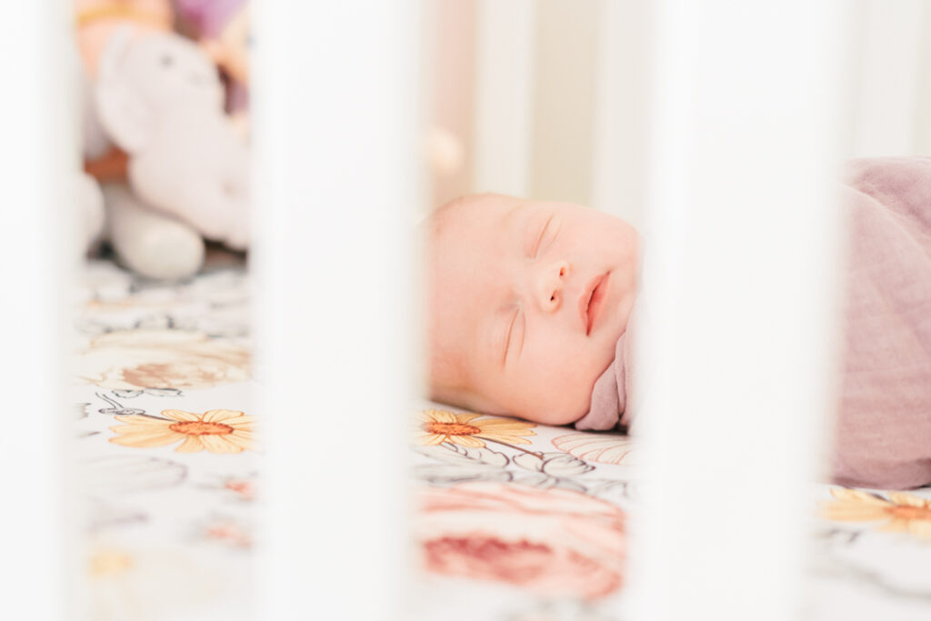 A newborn baby girl is peacefully sleeping in a white crib while swaddled in a mauve blanket. The photo is captured through the crib bars by an Austin newborn photographer during a lifestyle newborn session in Round Rock, TX. 