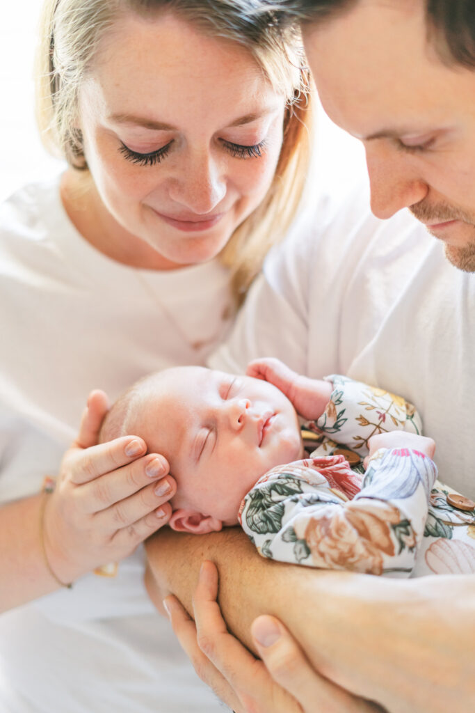 A couple lovingly cradles a sleeping baby in their arms, captured by an Austin Newborn Photographer. The baby, dressed in a natural floral outfit, is the center of their adoring smiles. This lifestyle newborn scene conveys warmth and tenderness.