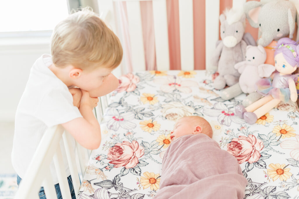 A little boy with blond hair is leaning over the front side of a white crib in a girl nursery filled with natural light coming from the window to his side. The boy looks down at his newborn sister, peacefully sleeping in her crib on a floral fitted shit, swaddled in a mauve blanket. To the right side of the crib are four little stuffed animals. The moment is captured during a lifestyle newborn session in Round Rock, TX. 