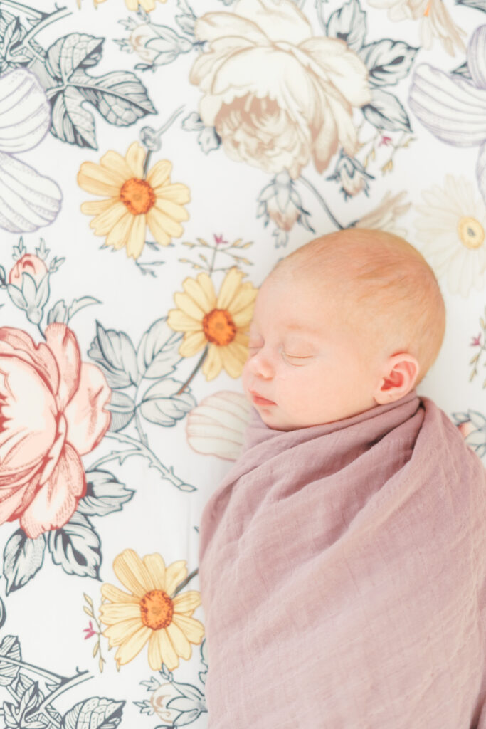 A newborn baby girl swaddled in a mauve muslin blanket is peacefully sleeping on a floral sheet. The background is light and airy for this lifestyle newborn session in Round Rock, TX.