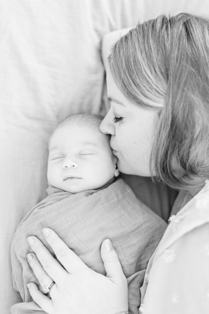 A black and white image captures a mother gently kissing her newborn, wrapped in a soft blanket. Her hand rests tenderly on the baby's side, conveying love and warmth, reminiscent of those cherished by an Austin newborn photographer.