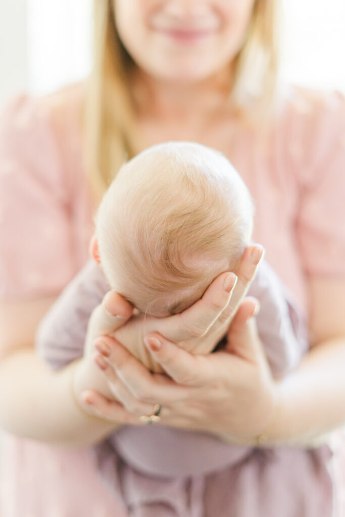 A mother in the blurred background holds her newborn baby's head in both her hands. The focus is on the back of the newborn's head and her beautiful blond hair. The mother wears a pink dress while the newborn is swaddled in a mauve muslin blanket. The background is filled with natural light, beautifully captured by an Austin newborn photographer.