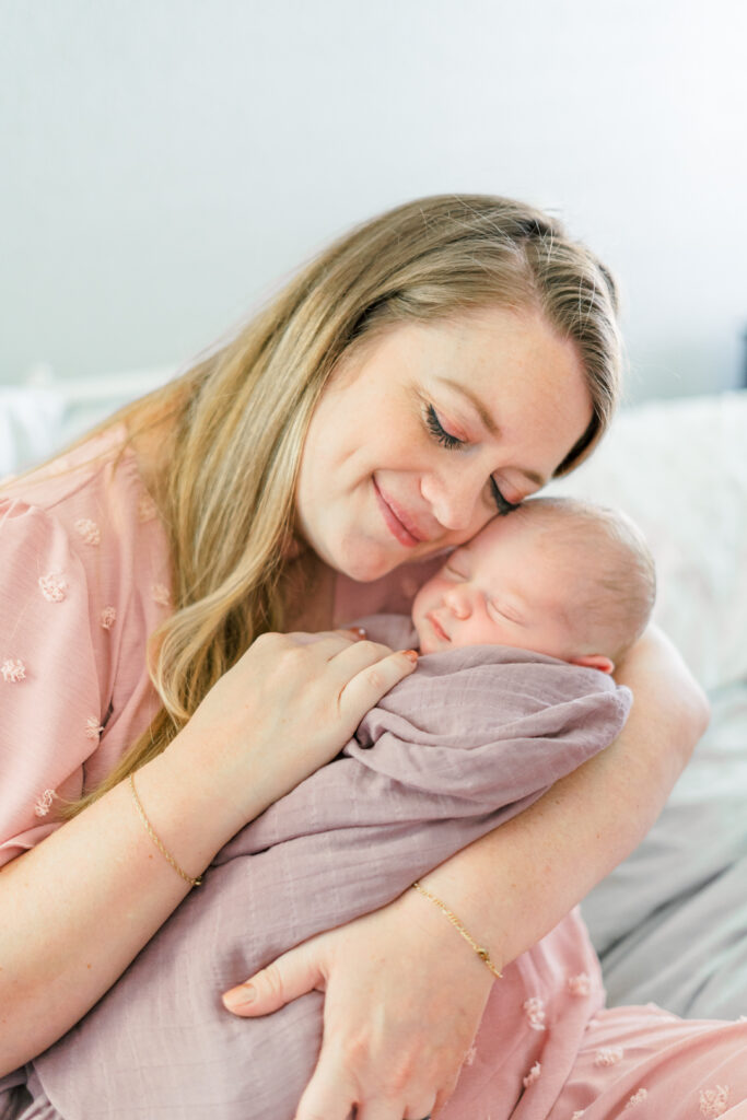 A woman in a pink dress is lovingly cradling her newborn wrapped in a mauve blanket. She sits on a bed with light-colored sheets, smiling softly with her eyes closed. The scene conveys warmth and tenderness, creating a light and airy atmosphere.