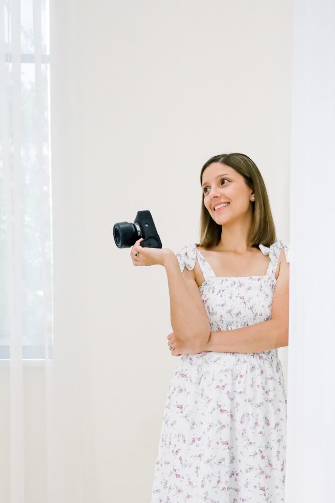 An Austin family photographer in a long white floral dress leans against an indoor white wall while holding up her camera in her right hand. She looks up and smiles. Light peeks through a window covered by a light curtain in the background. 