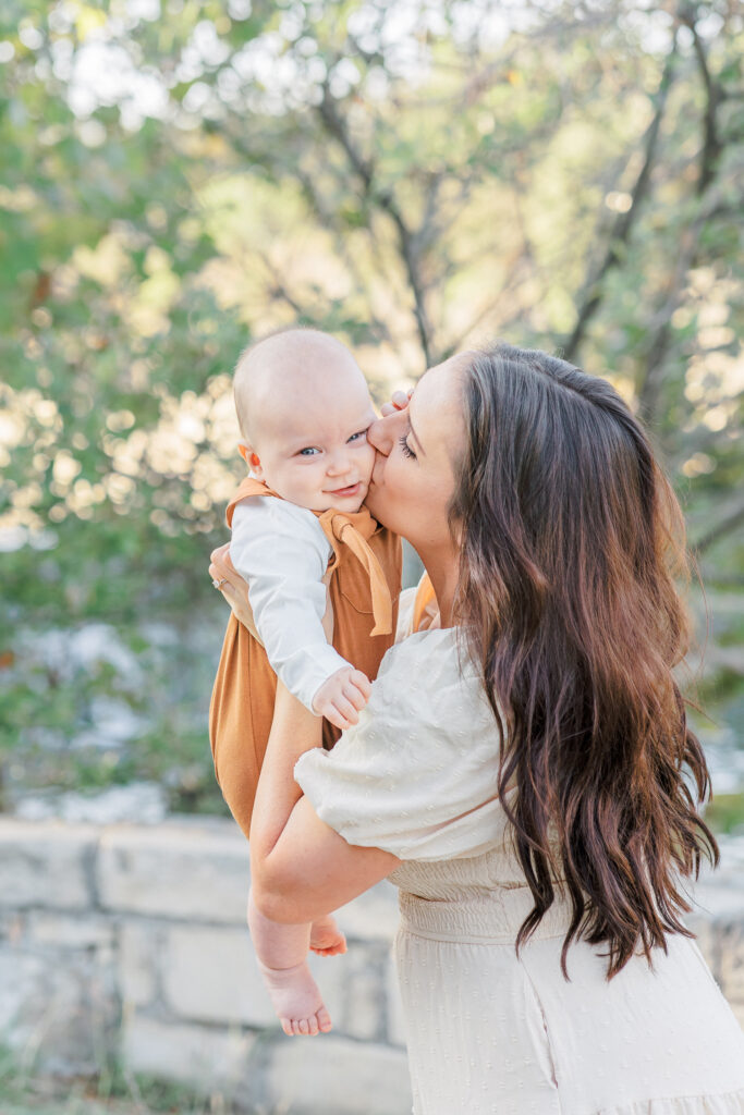 A mother stands holding her baby in front of her. She gives the baby a kiss on the cheek while the baby looks at the camera smiling. The baby wears a burnt orange jumper and the mother wears a long tan dress in this fall family photo captured by an Austin Family Photographer.