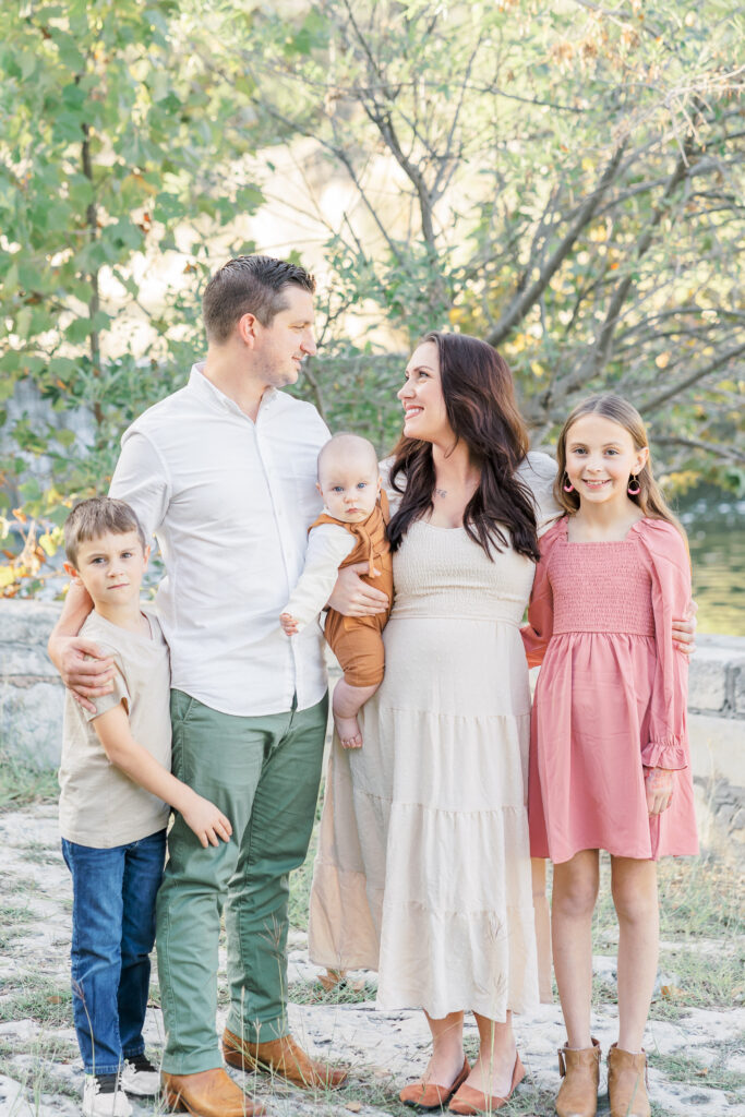 A family of 5 stands in a front of a lake at Blue Hole Park in Georgetown, TX. The mother and father look at each other smiling. The mother holds their baby in between them. The girl is next to her mother and the boy is next to his father. The photo is captured by an Austin Family Photographer