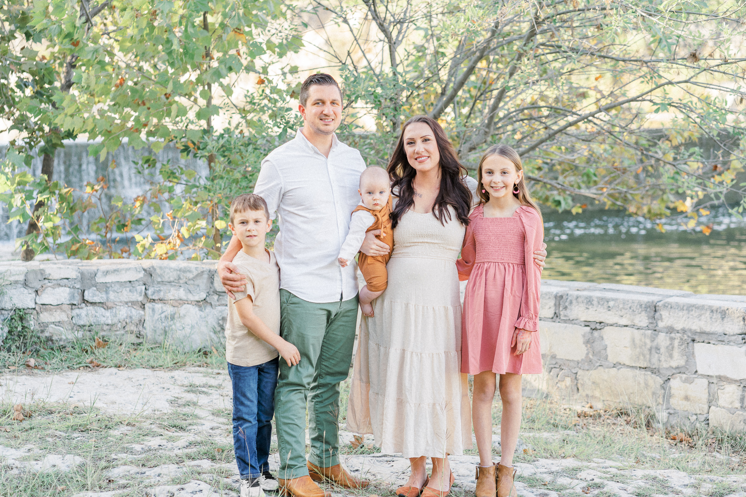 A family of five stands in front of a lake at Blue Hole in Georgetown, TX. The mother and father stands in the middle, with a baby on the mother's right hip, while a boy stands next to his father and a girl stands next to her mother. The photo is captured by an Austin Family Photographer.