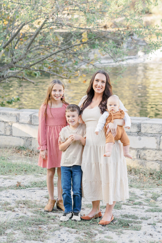 A mother stands in front of a lake at Blue Hole in Georgetown, TX with her three children, a girl, a boy, and a baby. She holds the baby in her arms and wraps her other arm around the boy. The girl, in a dusty rose dress, stands next to her brother