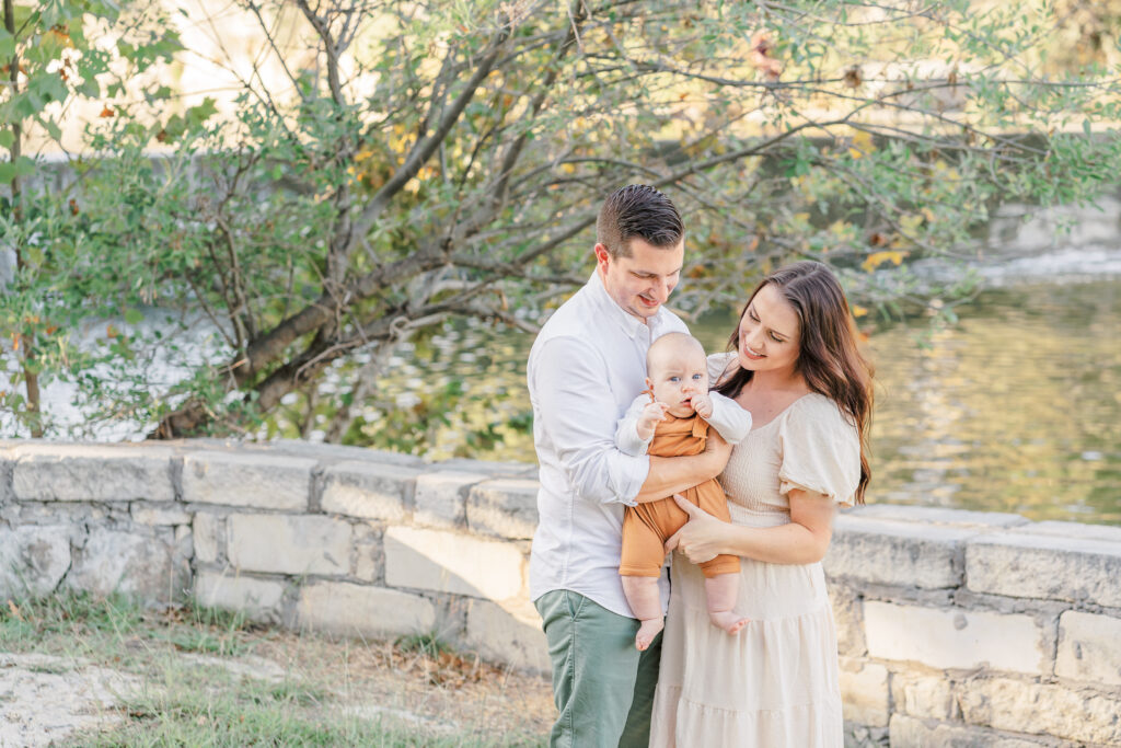 A mother and father stand in front of a lake in Blue Hole in Georgetown, TX. They hold their baby in between them and look at him smiling. The photo is captured by and Austin Family Photographer.