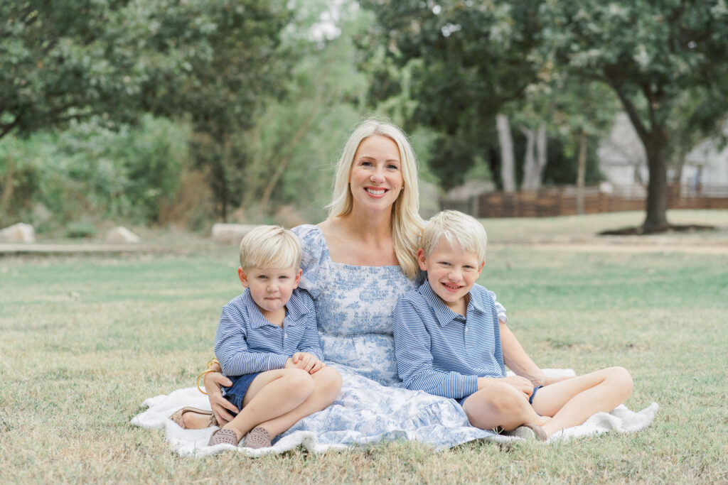 A mother and her two young sons sit on a white blanket at Katherine Fleischer Park in Austin, TX. Each boy is sitting next to their mother while they look and smile at the camera. The mother wears a long white and blue dress. The two young boys wear navy blue matching outfits in this image captured by an Austin Family Photographer.