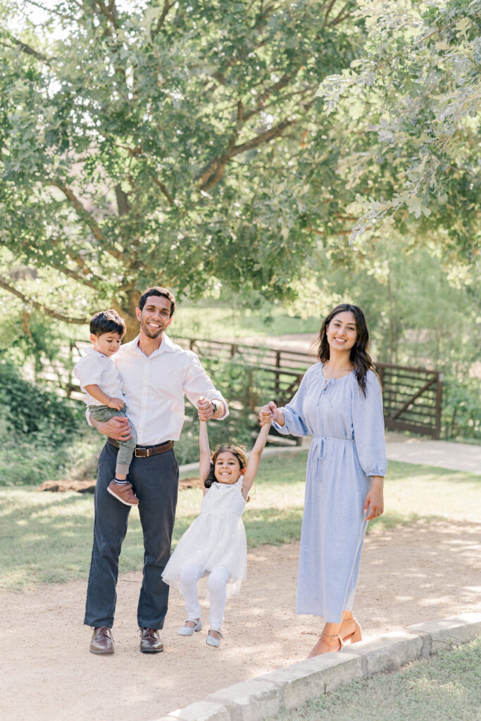 A family of four walks at Katherine Fleischer Park in Austin, TX. The dad holds the young toddler in his arms while the little girl holds her parents hands in the middle. The parents swing the little girl in between them. The family is laughing and having fun in this authentic shot captured by an Austin Family Photographer.