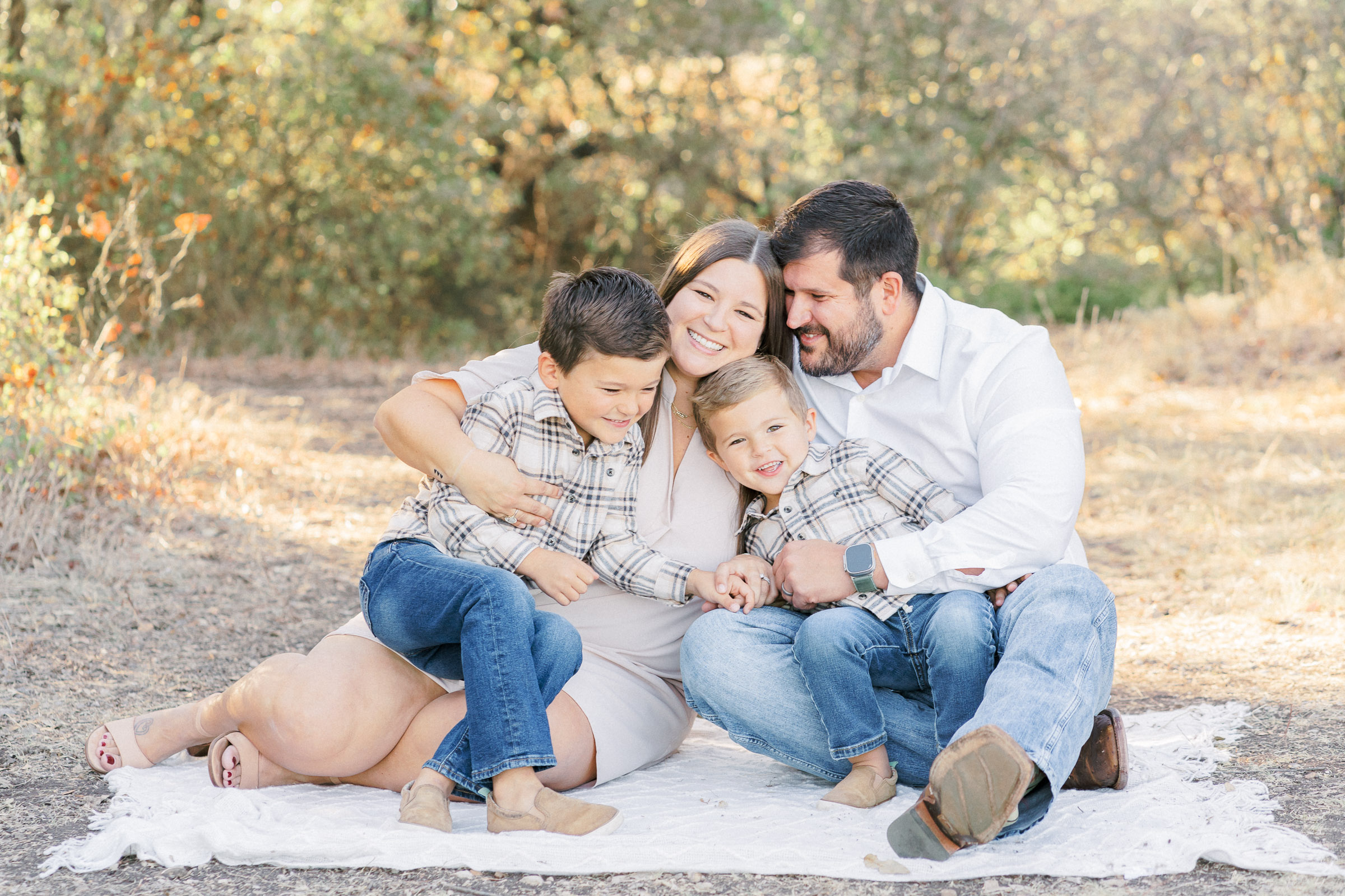 A family of four sits on a blanket at Brushy Creek Lake Park in Cedar Park, TX. The family hugs each other. The two young boys wear matching outfits including blue jeans and plaid collar shirts. The family laughs together in this beautiful shot captured by an Austin Family Photographer