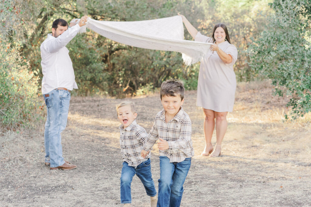 A mother and father hold up a white blanket while their two young boys, in matching plaid shirts, run underneath the blanket at Brushy Creek Lake Park in Cedar Park, TX. The family laughs and radiants joy in this tender shot captured by an Austin Family Photographer.