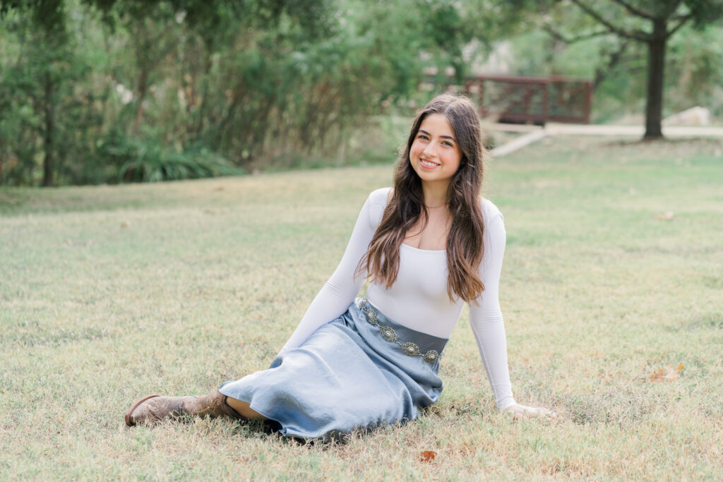 a young girl sits on the grass at Katherine Fleischer Park in Austin, TX. She wears a white shirt with a long blue skirt. She smiles in this authentic moment captured by an Austin Family Photographer.