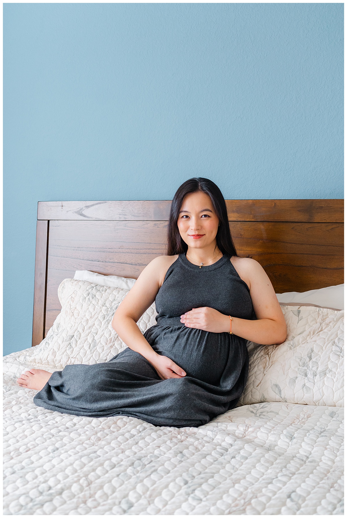 a pregnant woman in a long charcoal dress sits on a bed. She holds her belly with both hands and looks at the camera with a soft smile in this indoor maternity portrait.