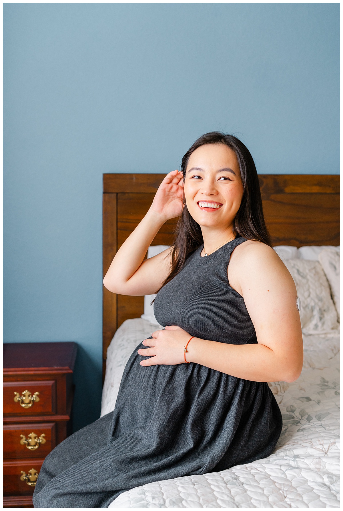 a pregnant woman dressed in a long charcoal dress sits at the edge of a bed. She has one hand on her belly and pushes her hair away with the other hand while smiling during her maternity photoshoot at home.