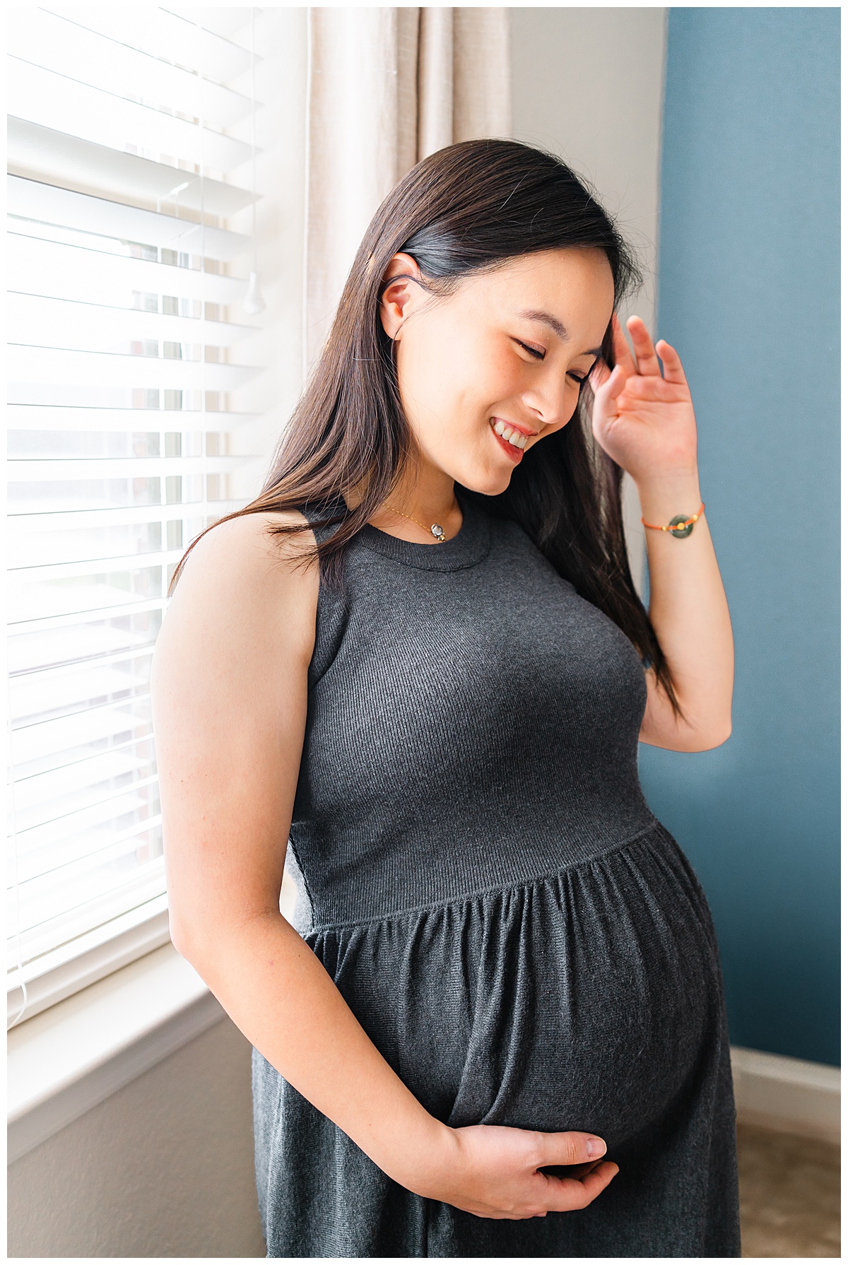 a pregnant woman dressed in a long charcoal dress stands against a window. She holds her belly with one hand and pushes her hair away with the other hand. She looks down with a soft smile on her face in this photo taken during a maternity photoshoot at home.