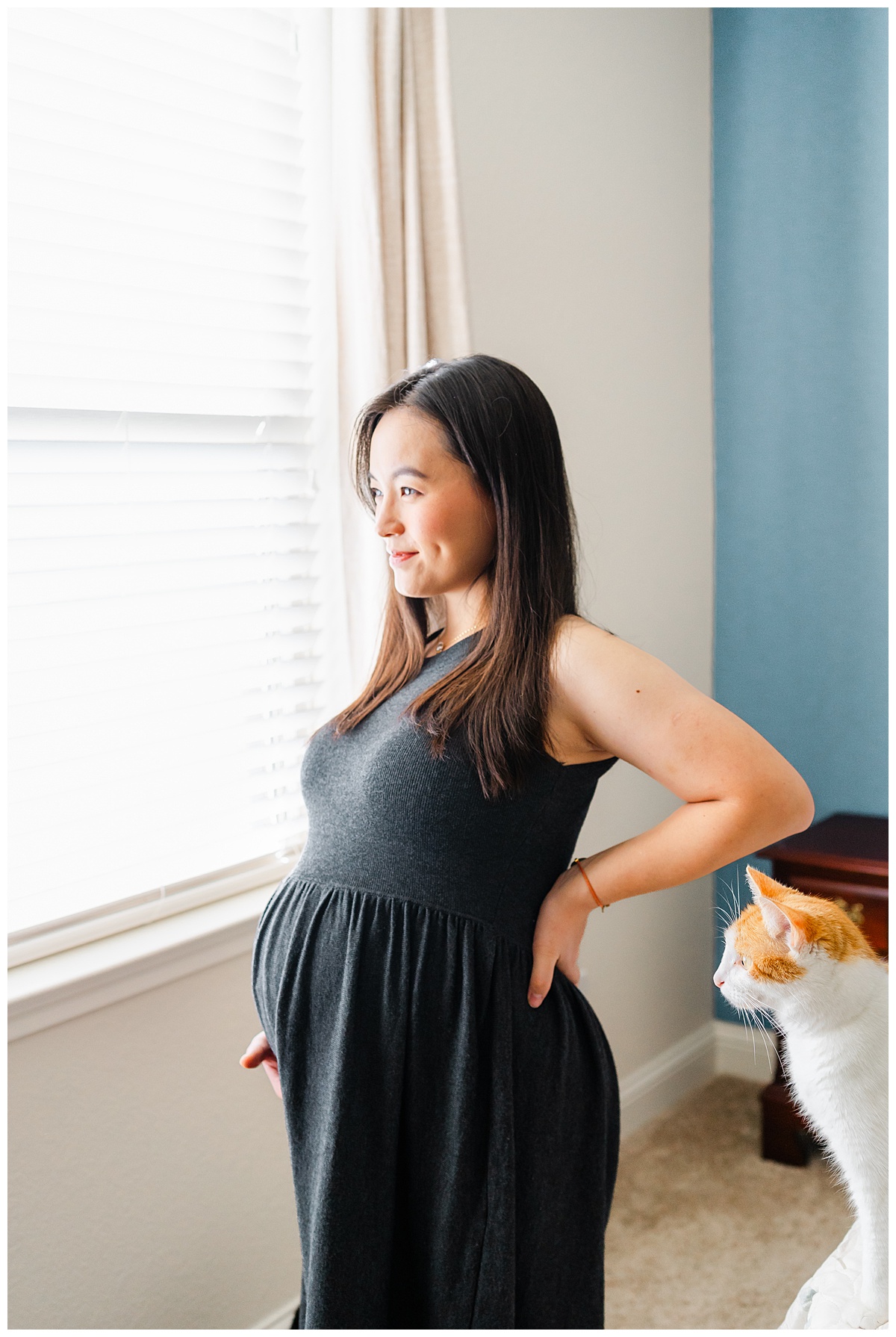 a pregnant woman dressed in a long charcoal dress stands in front of a window. On the bed next to her is a white and orange cat. They both look away at the window in this cute shot taken by a maternity photographer in Austin, TX.