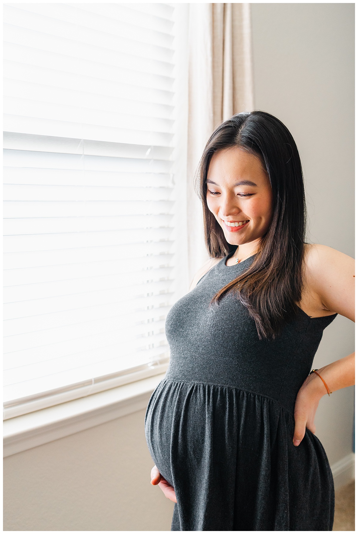 a pregnant woman dressed in a long charcoal dress stands in front of a window. She looks down at her belly and smiles in this maternity photo.