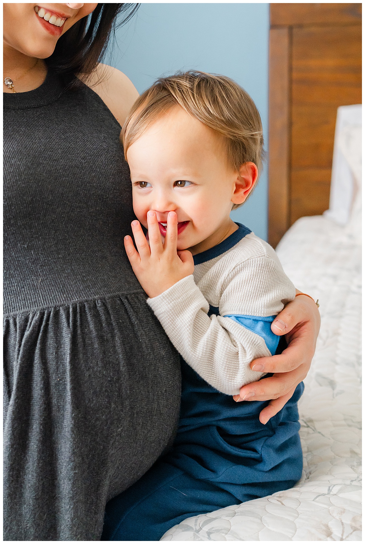a little boy snuggles with his pregnant mother while sitting on the edge of a bed during this indoor maternity photoshoot.
