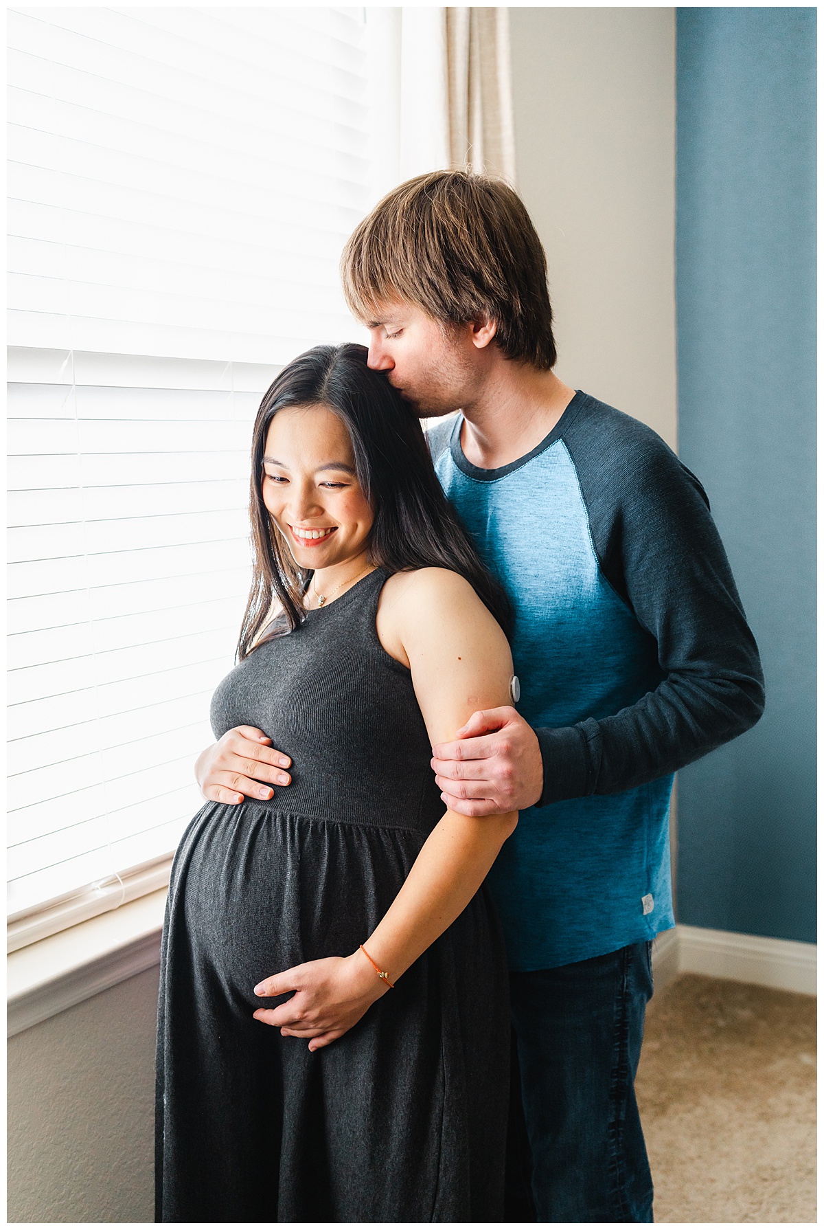 a pregnant mother and her husband stand in front of a window. The husband stands behind the mother and kisses her on the back of her head. The mother looks down smiling in this sweet shot taken by a maternity photographer in Austin, TX.