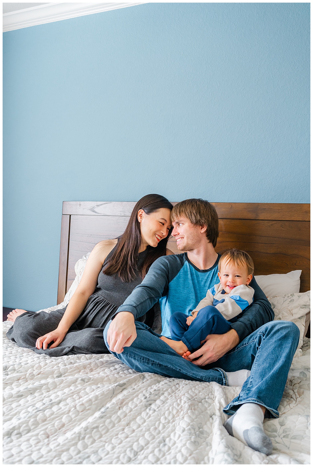 a family of three sits on a bed. The little boy sits in his father's lap and looks at the camera smiling. The parents have their foreheads together and smile at each other during this in home maternity photoshoot in Austin, TX.