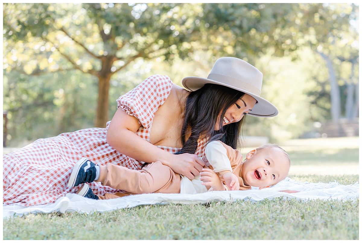 A mother dressed in a burnt orange gingham dress lay on a white blanket with her baby boy at Katherine Fleischer Park. She tickles her baby while he laughs during their sweet mommy and me photoshoot in Austin, TX.