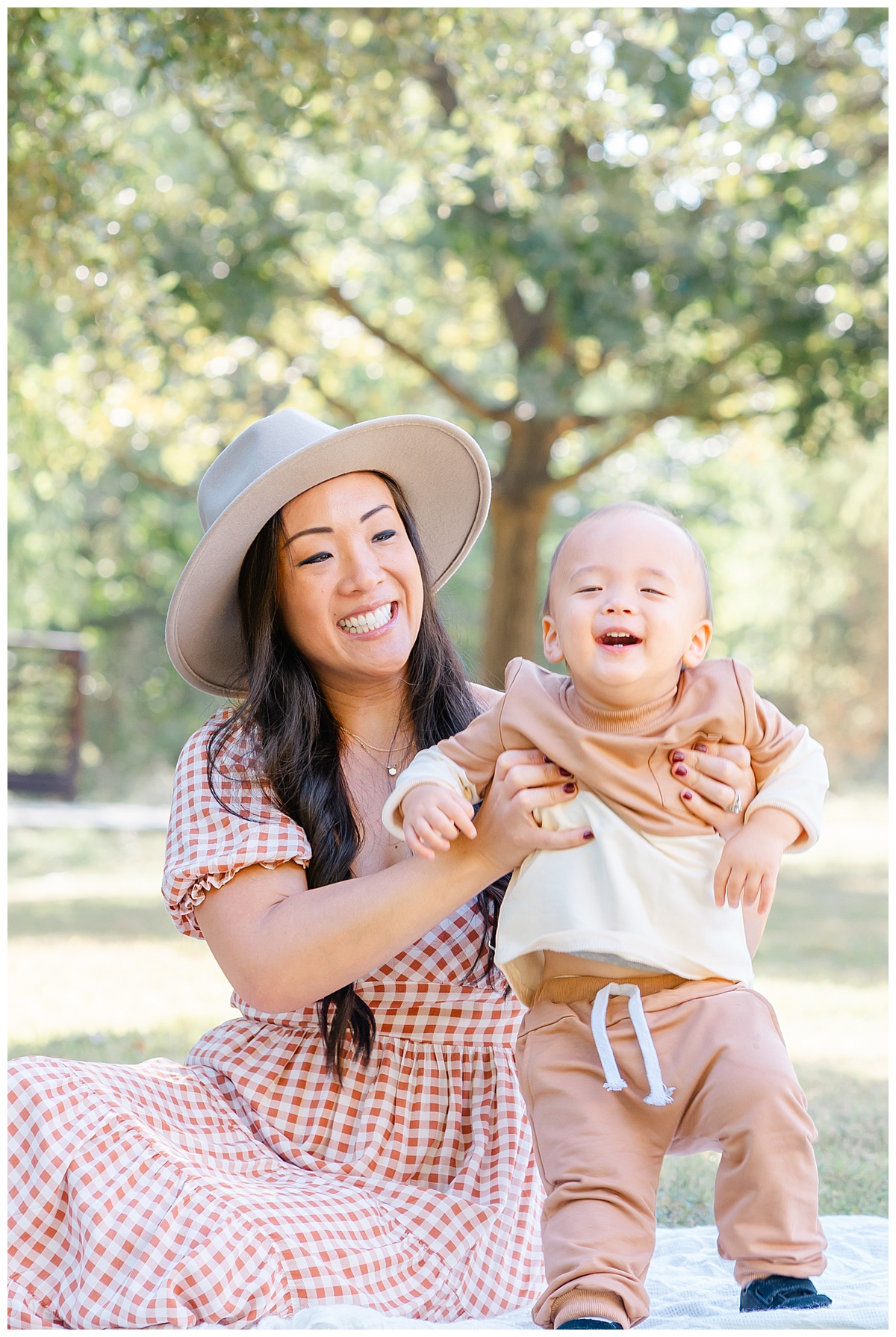 Austin Moms | A mother dressed in a burnt orange gingham midi dress and wearing a tan hat sits on a white blanket with her baby boy. She is ready to throw her baby in the air while he is joyfully laughing during their mommy and me photoshoot.