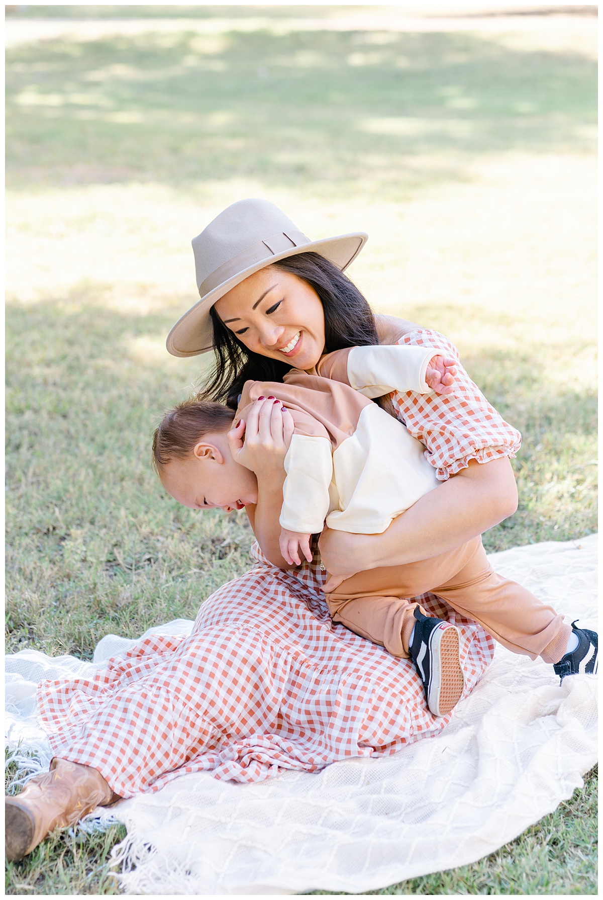 Austin Moms | A mother dressed in a burnt orange gingham dress and wearing a tan hat sits on a white blanket with her baby boy at Katherine Fleischer Park.