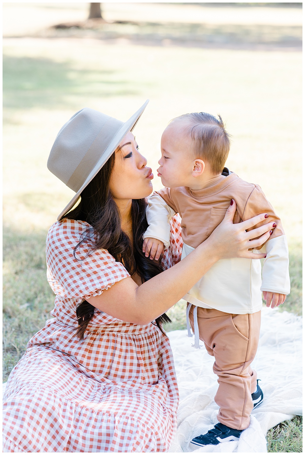 Austin Moms | A mother sits on a white blanket. She kisses her baby boy standing next to her.