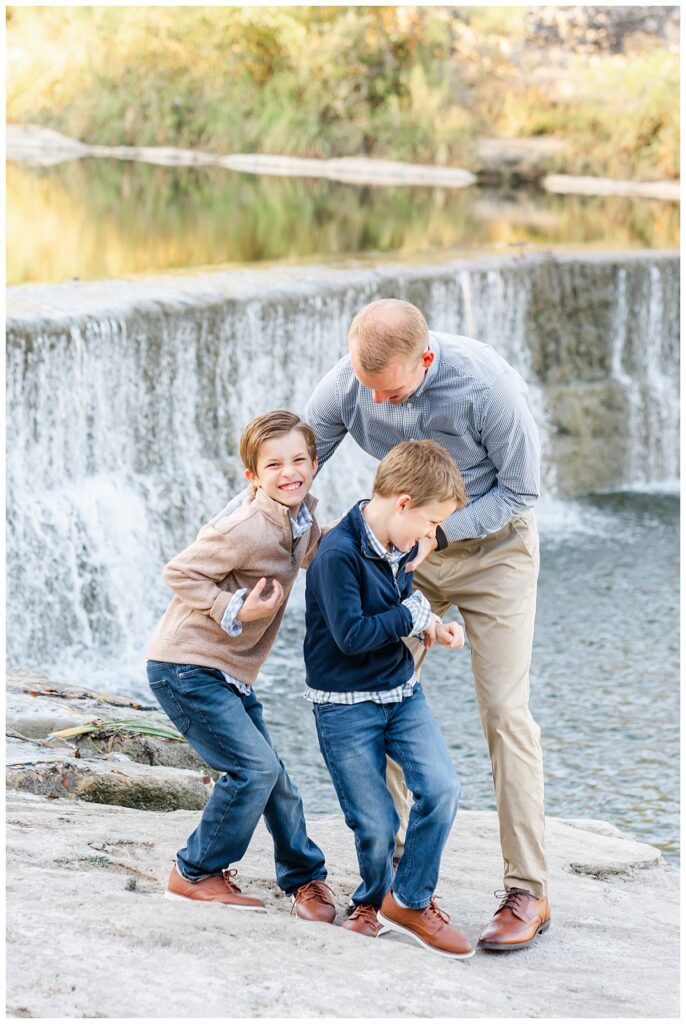A father and his two young boys play a tickle fight in front of a waterfall at Blue Hole Park. They all laugh together in this candid shot taken by a Georgetown Texas Photographer.