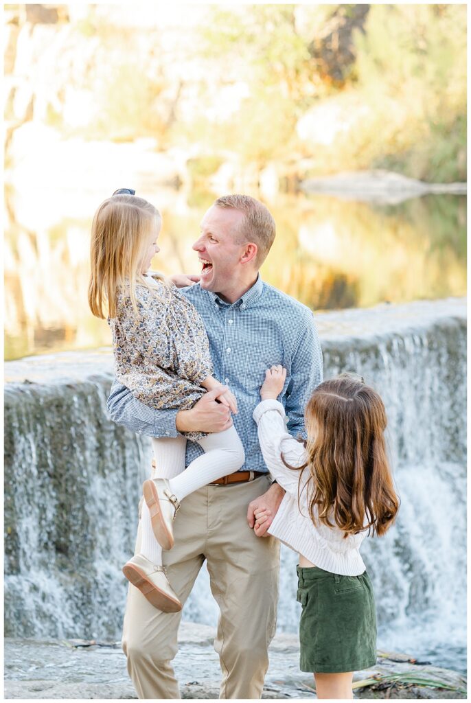 A father and his two young daughters stand in front of a waterfall at Blue Hole Park. The father holds his youngest daughter and tickles her while she laughs. The oldest daughter stands by her father and tickles him in this family shot captured by a Georgetown Texas Photographer.