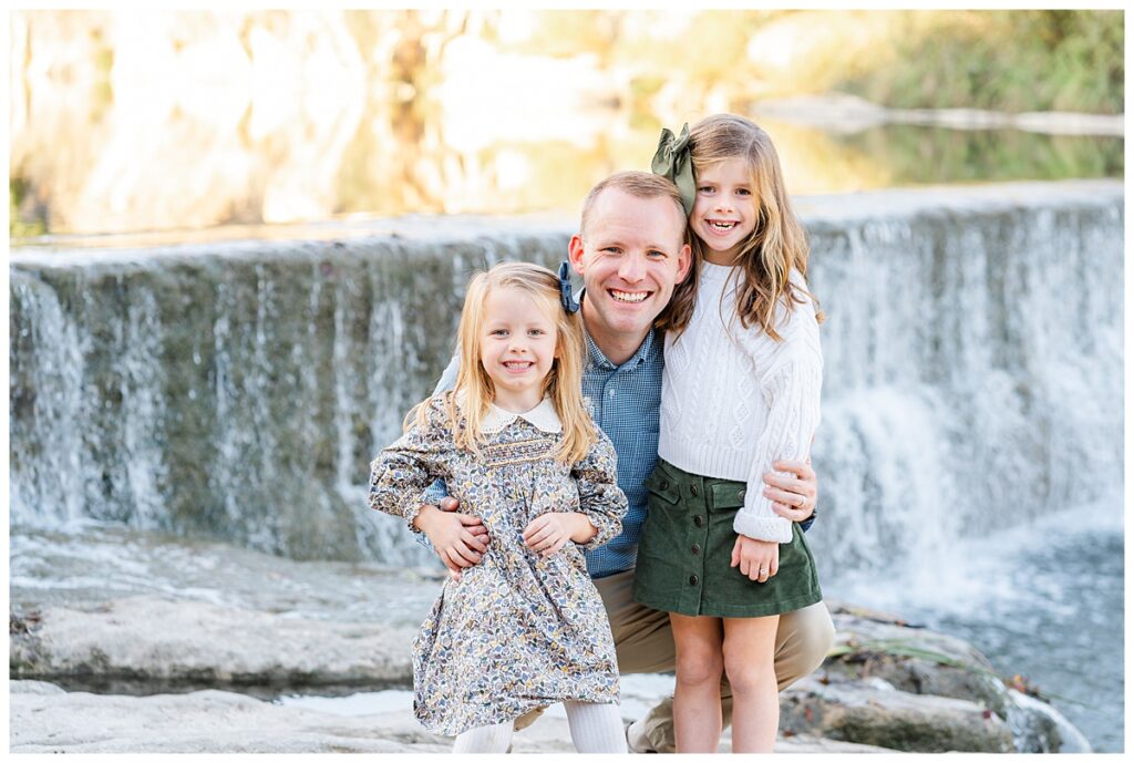 A father and his young daughters sit in front of a waterfall at Blue Hole Park. They all look at the camera smiling in this posed photo taken by a Georgetown Texas Photographer.