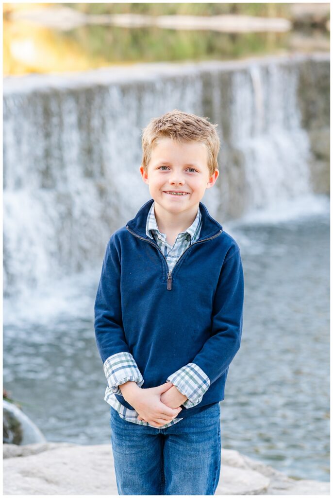 A young boy wearing braces poses and smiles in front of a waterfall at Blue Hole Park for family photos captured by a Georgetown Texas Photographer.