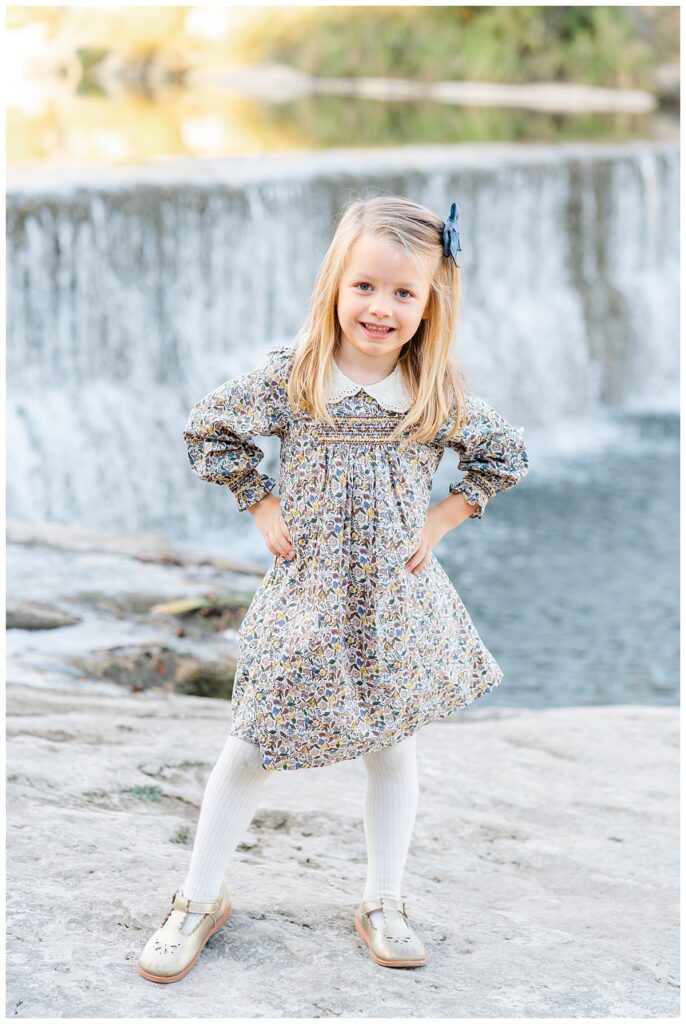 A young blond girl in a floral dress with a white peter pan collar poses with her hands on her hips, in front of a waterfall at Blue Hole Park. She smiles at the camera in this sweet portrait captured by a Georgetown Texas Photographer.