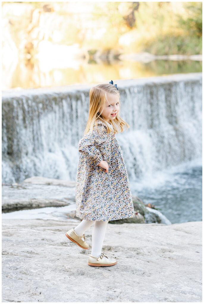A young blond girl in a floral dress with white tights twirls in front of a waterfall at blue hole park. She smiles and looks happy during her family photos taken by a Georgetown Texas Photographer.