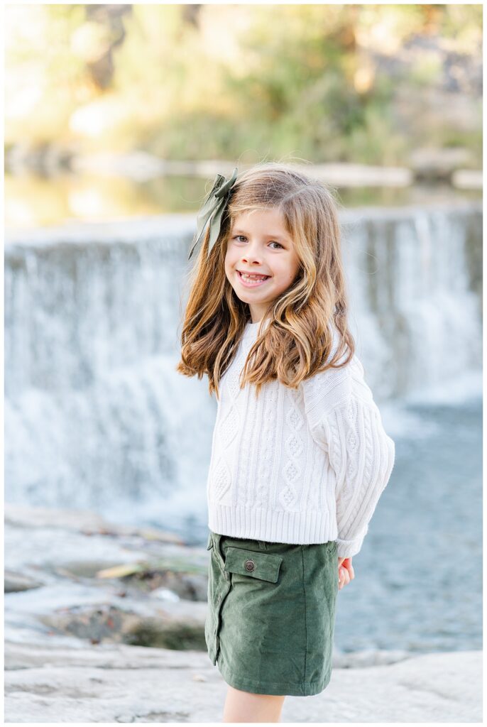 A young girl with long black hair dressed in a green skirt and while sweater stands in front of a waterfall at blue hole park. She poses with her hands behind her back and smiles at the camera in this posed portrait taken by a Georgetown Texas Photographer.