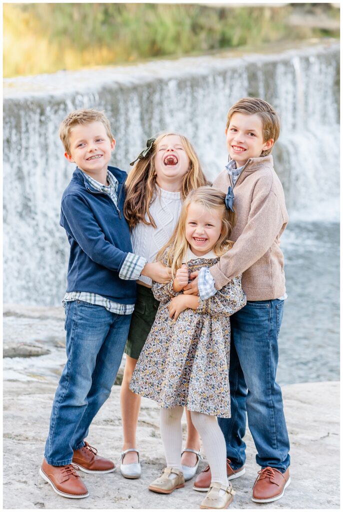 A group of four siblings including two girls and two boys stand in front of a waterfall at Blue Hole Park. They tickle each other and laugh in this candid moment captured by a Georgetown Texas Photographer.