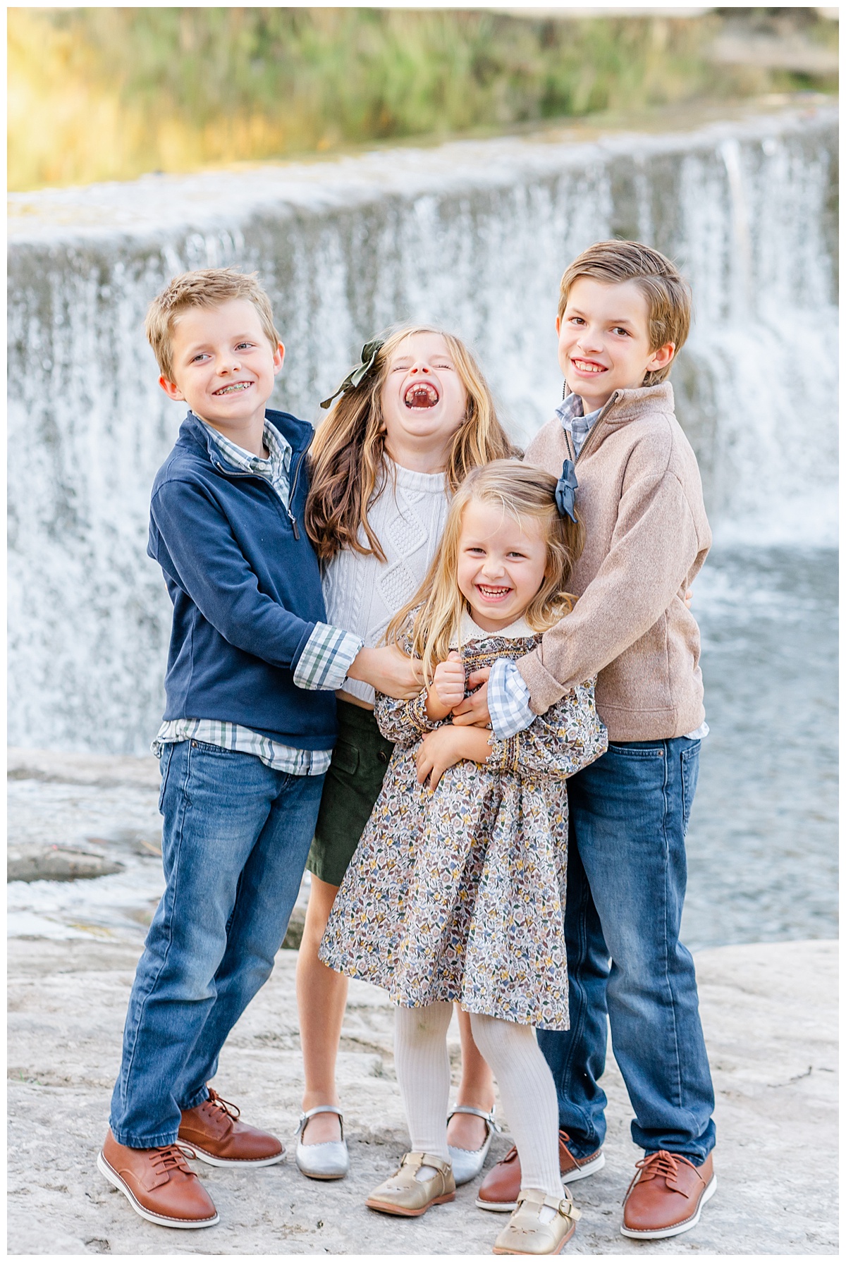 A group of four siblings including two girls and two boys stand in front of a waterfall at Blue Hole Park. They tickle each other and laugh in this candid moment captured by a Georgetown Texas Photographer.