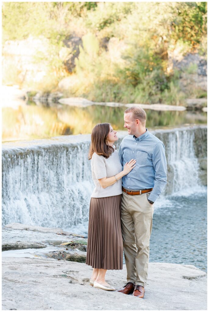 A couple stands in front of a waterfall at blue hole park. The woman wear a tan top and long brown skirts while her husband wears a light blue collar shirt and khaki pants. The woman is close to her husband with her hand on his chest. They face each other and look at each other with a soft smile in this sweet photo taken by a Georgetown Texas Photographer.