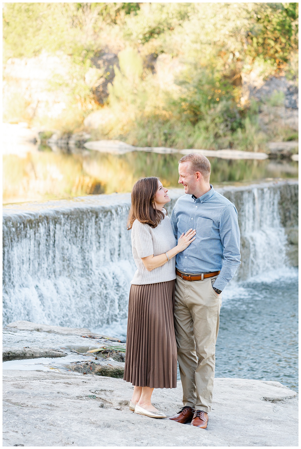 A couple stands in front of a waterfall at blue hole park. The woman wear a tan top and long brown skirts while her husband wears a light blue collar shirt and khaki pants. The woman is close to her husband with her hand on his chest. They face each other and look at each other with a soft smile in this sweet photo taken by a Georgetown Texas Photographer.