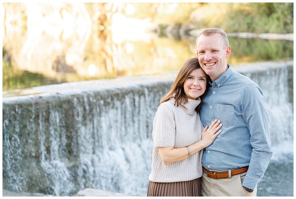 A couple stands in front of a waterfall at Blue Hole Part. The woman leans her head on the man's shoulder and places her hand on his chest. They both look at the camera and smile in this posed couples portrait captured by a Georgetown Texas Photographer.