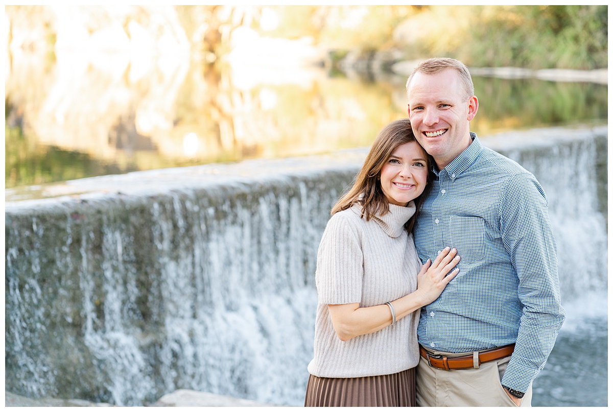 A couple stands in front of a waterfall at Blue Hole Part. The woman leans her head on the man's shoulder and places her hand on his chest. They both look at the camera and smile in this posed couples portrait captured by a Georgetown Texas Photographer.