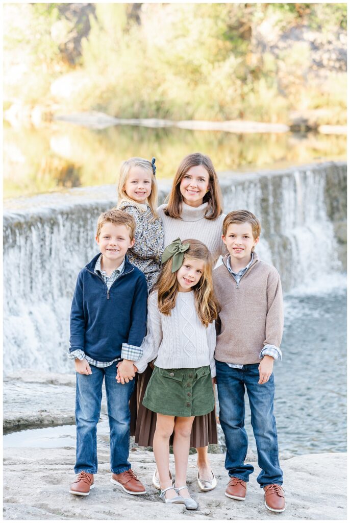 A mother and her four children stand in front of a waterfall at blue hole park. They pose and smile at the camera in this family photo taken by a Georgetown Texas Photographer